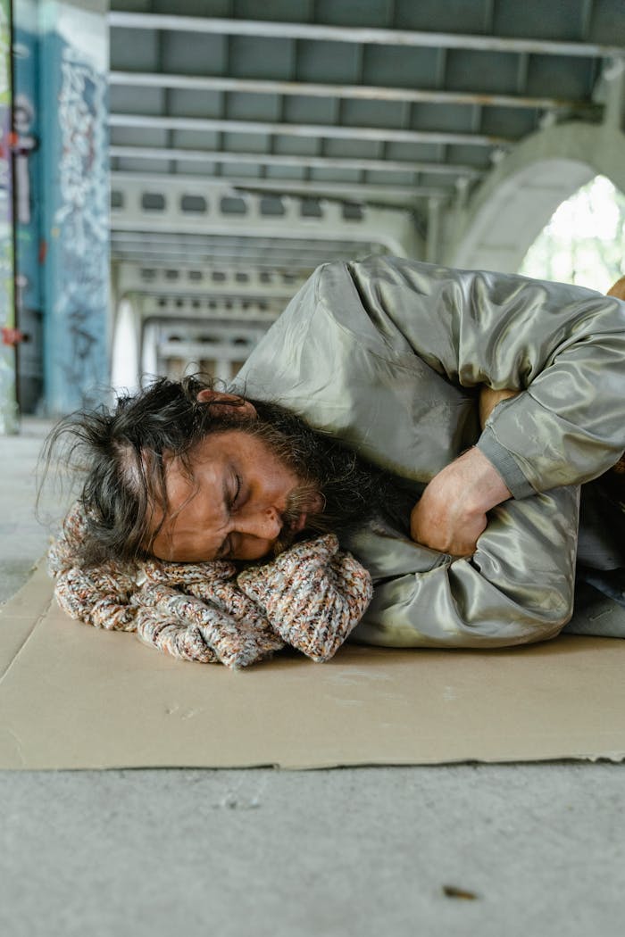 services-04 A homeless man with a beard is sleeping on a cardboard under a bridge, highlighting urban poverty.