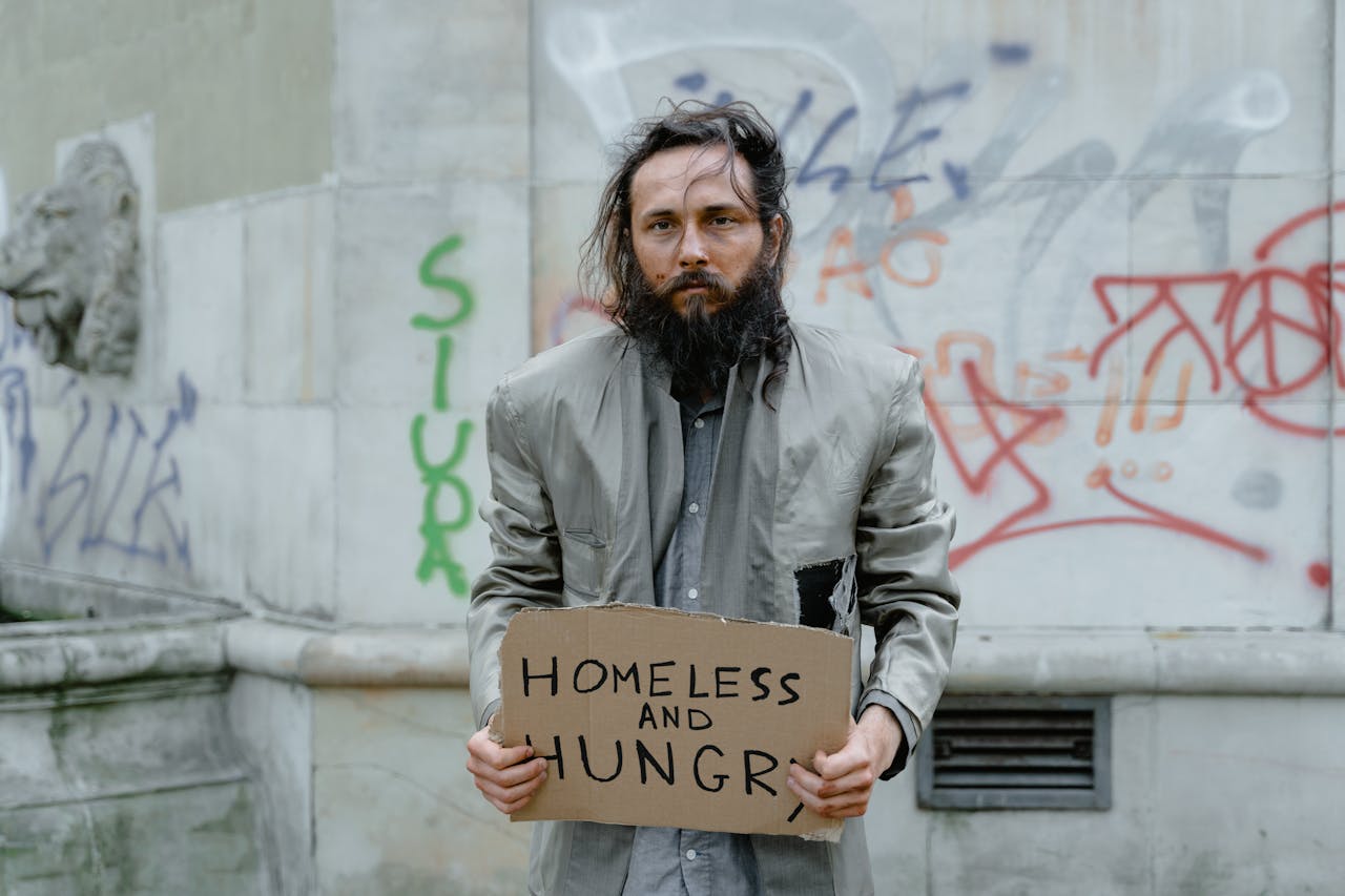 heros-img Homeless man standing on the street holding a sign, with graffiti in the background.