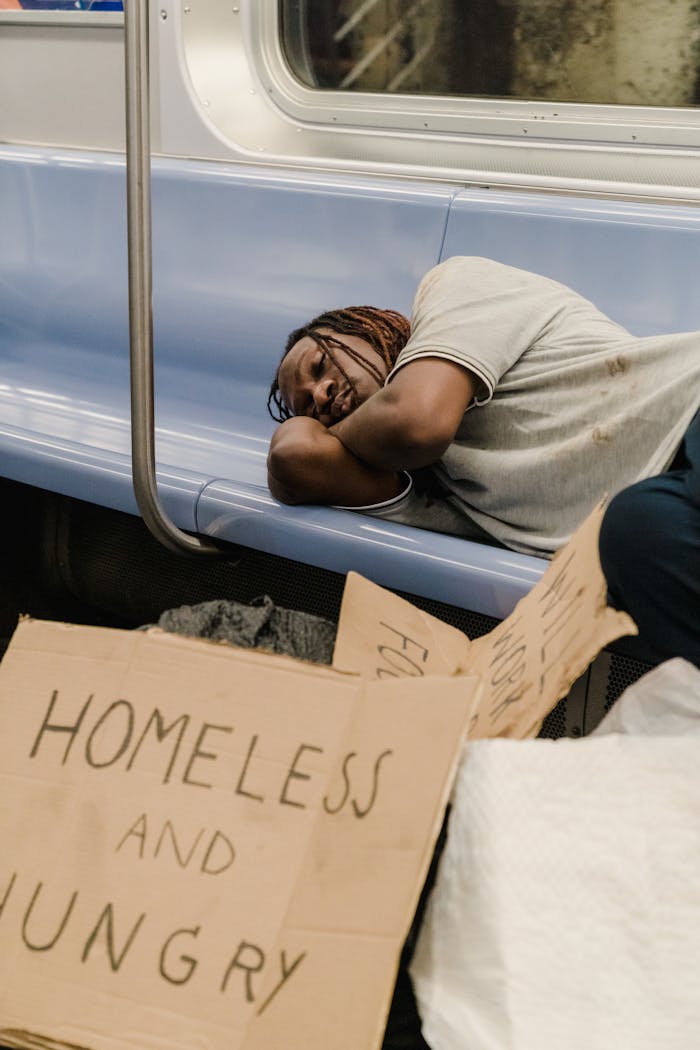 services-03 An African American homeless man is sleeping on a subway bench next to cardboard signs.