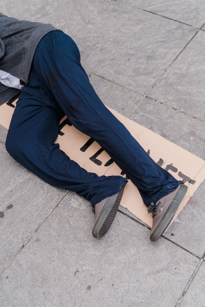 about-us A person in blue pants lies on a sidewalk with a cardboard sign, symbolizing homelessness.